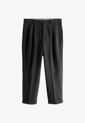 Junger Mann mit lockigem Haar, der einen schwarzen Blazer, ein schwarzes Hemd, passende schwarze Hose und schwarze Slipper trägt und vor einer vertäfelten Wand steht.