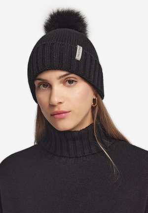 Ung kvinde iført sort strikket hue med pompon og Falcone-mærke, sort rullekravetrøje og gylden ørering, neutralt baggrund.