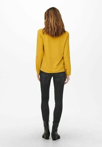 Haut à manches longues jaune moutarde avec poignets à boutons, accompagné d'un jean skinny noir et de bottines, vu de dos sur fond blanc.
