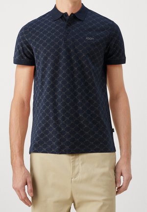 Mężczyzna ubrany w granatową koszulkę polo w wzory z logo "JOOP!" na piersi i beżowe spodnie, stoi na tle jednolitego tła.