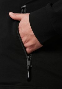 Sudadera con cremallera negra que presenta un bolsillo frontal, una tela suave y un tirador de cremallera texturizado con el logo "CAMP DAVID". Vista en primer plano de una mano en el bolsillo.