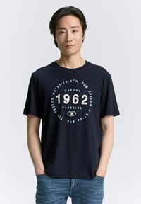 Camiseta de algodón azul marino de manga corta con un diseño gráfico redondo en blanco y azul claro, que muestra '1962' y el logo de la marca.