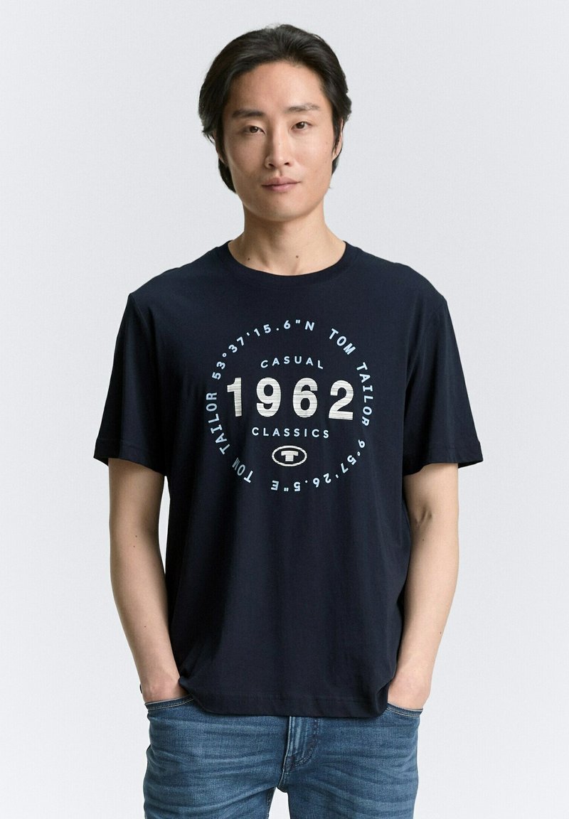 Camiseta de algodón azul marino de manga corta con un diseño gráfico redondo en blanco y azul claro, que muestra '1962' y el logo de la marca.
