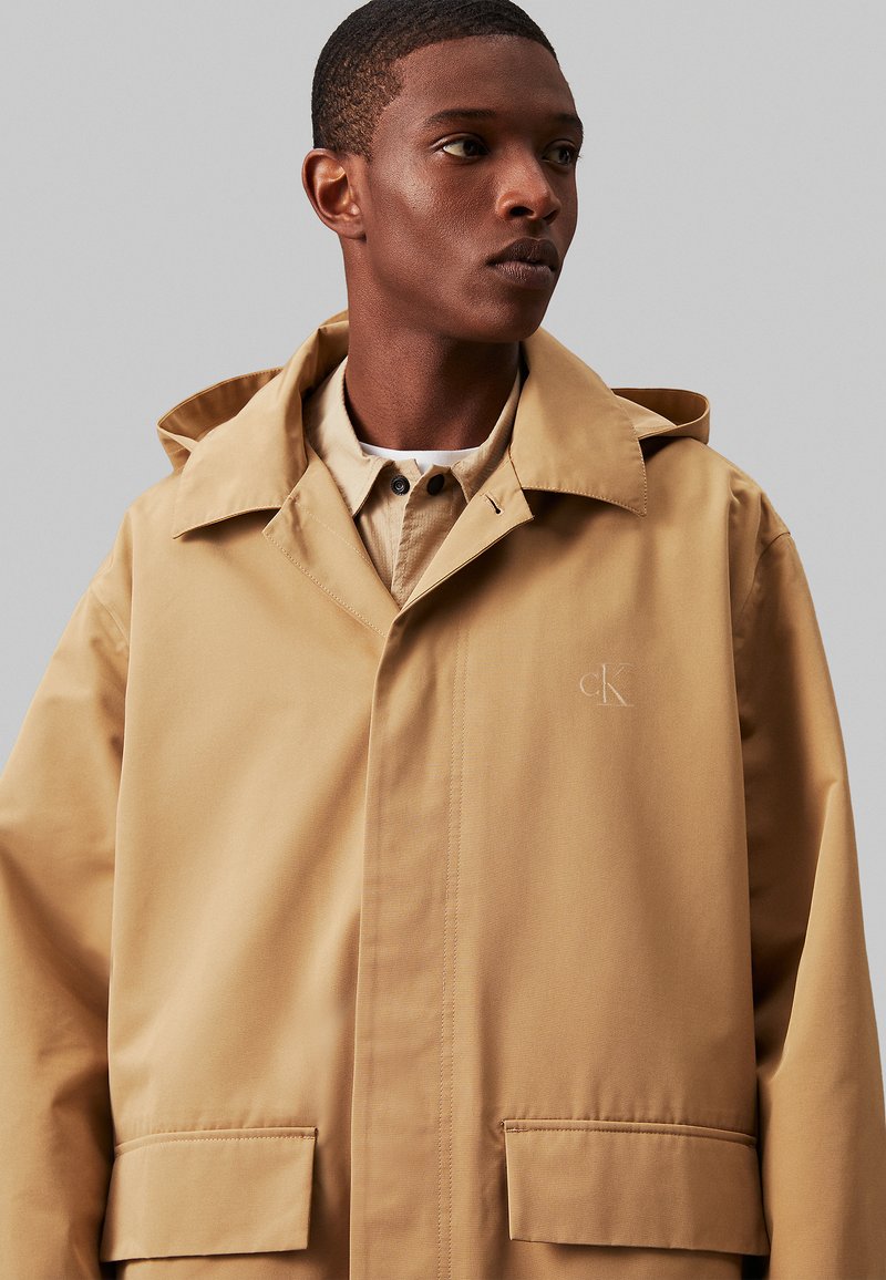 Beżowa kurtka wodoodporna z dużym kapturem, kieszeniami z przodu i subtelnym logo na klatce piersiowej. Gładki, trwały materiał o prostym designie.