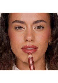 Lip crayon in soft red with a creamy texture, held near lips. Model has medium brown skin, dark curly hair, and natural makeup. Silver hoop earrings.