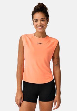 Glunderende vrouw met krullend haar in een perzikkleurige mouwloze sporttop en zwarte korte broek, die een vossen-tatoeage op haar linkerarm toont.