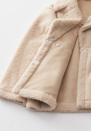Veste en fausse fourrure beige avec une texture douce, un col à revers et des boutons-pression métalliques. Doublure intérieure lisse et beige.
