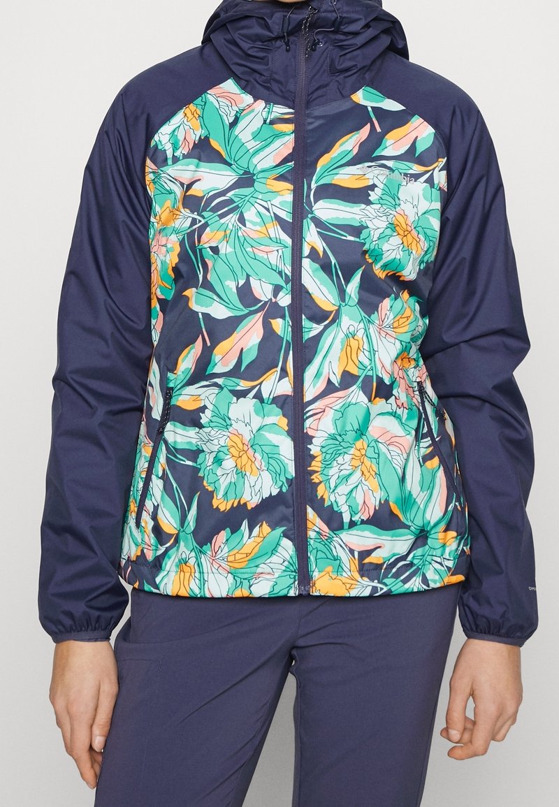 Veste bleu marine avec fermeture éclair devant et capuche, panneaux avant ornés d’un grand motif floral vert, jaune et rose, portée avec un pantalon bleu marine assorti.