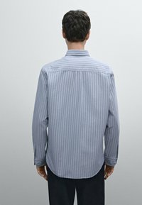 Koszula z długim rękawem w jasnoniebieskim kolorze z białymi pionowymi paskami. Posiada kołnierzyk, zapięcia na guziki przy mankietach oraz zaokrąglony dół. Wykonana z bawełnianej tkaniny.