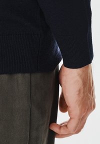 Granatowy sweter wełniany z ściągaczami, noszony na oliwkowych spodniach; ręka oparta na boku. Miękka tekstura i dopasowany fason.