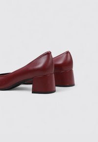 Chaussures à talons bordeaux avec une finition brillante, présentant un design à bout arrondi et un talon bloc. Texture en cuir lisse, style minimaliste.