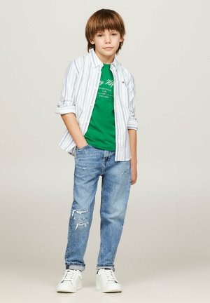 Ragazzo che indossa una camicia bianca a righe con bottoni sopra una T-shirt verde, abbinata a jeans blu strappati e sneakers bianche.