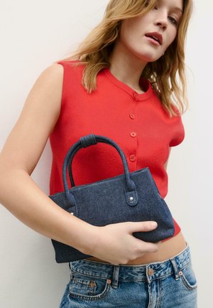 Donna che indossa un top rosso senza maniche con bottoni e jeans blu tiene una piccola borsa in denim blu scuro contro uno sfondo bianco.