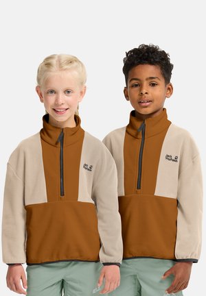 Deux enfants portant des vestes polaires à fermeture éclair Jack Wolfskin beige et marron, ainsi que des pantalons vert clair, debout côte à côte devant un fond blanc.