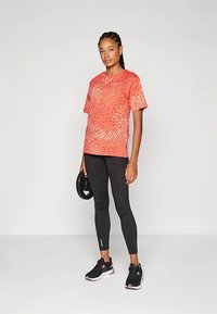 Raudonos ir oranžinės spalvos su raštais trumpomis rankovėmis marškinėliai, derinami su juodomis leginsėmis ir juodais sportiniais batais; marškinėliai turi bangų dizainą ir laisvą siluetą.