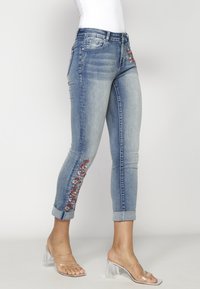 Hellblaue Jeans aus Denim mit floralem Stickerei in Rot und Weiß entlang der Seite, schlanke Passform und umgeschlagene Säume. Klare Sandalen mit Blockabsatz.