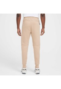 Pantaloni jogger beige realizzati in tessuto morbido, caratterizzati da un design affusolato, una vita elastica e due tasche laterali. Dettagli decorativi includono cuciture.