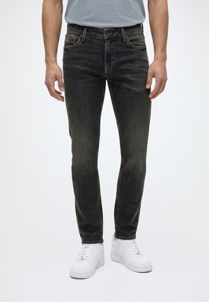 Homme portant un jean slim noir délavé et des baskets blanches, debout devant un fond uni.