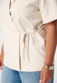 Haut wrap beige en tissu lisse, doté de manches courtes, de détails à nouer à la taille, associé à un jean bleu clair.