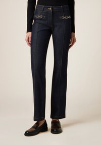 Pantalones de denim azul oscuro con pierna recta, destacados por distintivos acentos de metal en la cintura y detalles de costura amarilla en contraste.