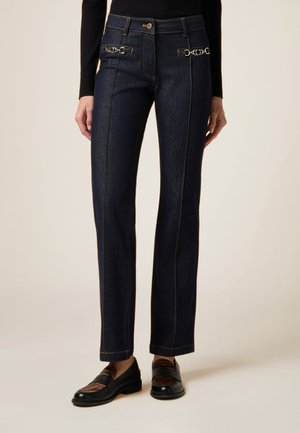 Pantalones de denim azul oscuro con pierna recta, destacados por distintivos acentos de metal en la cintura y detalles de costura amarilla en contraste.