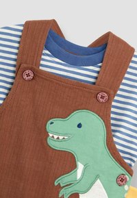 Brune kordfløjls overalls med dino-patch, parret med en blå og hvid stribet skjorte. Har pink knapper og ribbet tekstur.