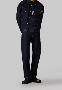 Mörk denimjacka med silverknappar, kombinerad med mörka denimjeans och svarta skor. Jackan har en klassisk design och synliga sömmar.