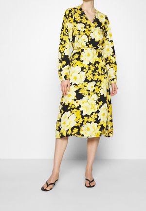 Femme portant une robe midi portefeuille noire avec un grand imprimé floral jaune et blanc, manches longues, et des sandales noires à bout ouvert.