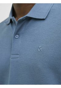 Polo majica u svijetloplavoj boji, od teksturiranog materijala. Ima ovratnik, dva dugmeta i mali izvezeni logo na prsima.