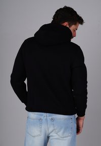 Black pullover hoodie with a drawstring hood, ribbed cuffs, and hem, paired with light blue jeans featuring back pockets.