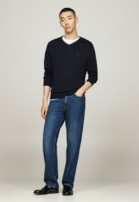 Marineblauer V-Ausschnitt-Pullover, weißes Unterhemd, blaue Jeans und schwarze Schuhe. Der Pullover hat gerippte Bündchen und ein kleines Logo auf der Brust. Lässige Passform.
