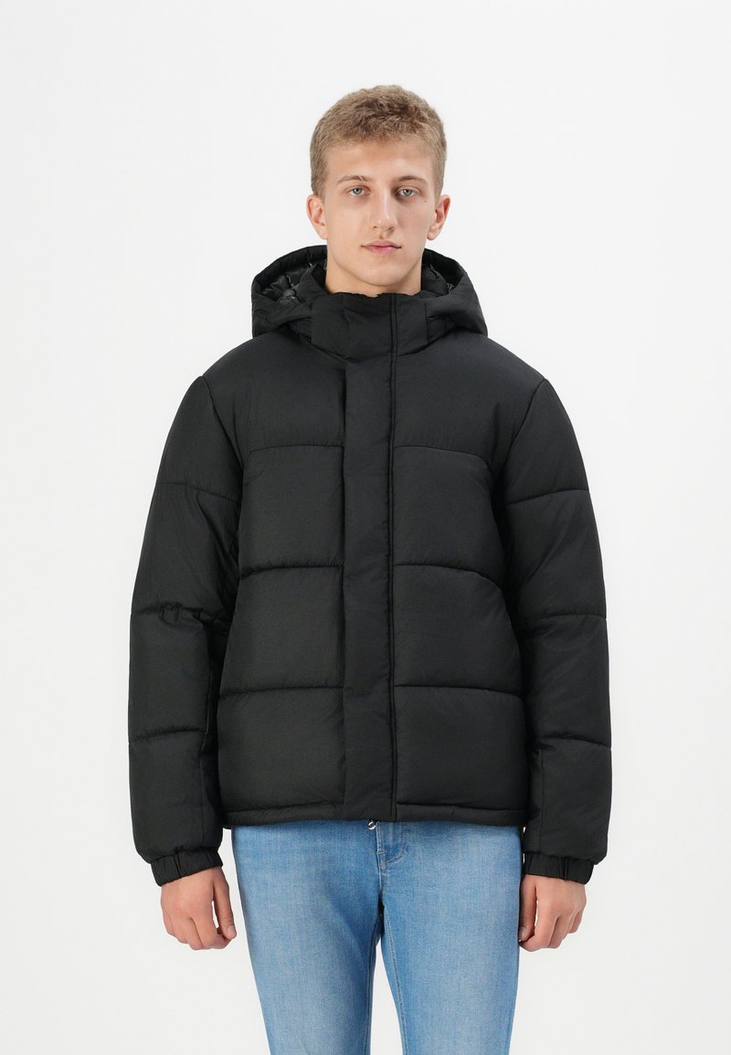 Schwarze Steppjacke mit Kapuze, featuring quadratischen gesteppten Abschnitten und elastischen Bündchen. Kombiniert mit hellblauen Jeans. Schlichtes und funktionales Design.