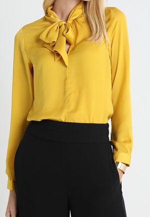Kobieta ubrana w musztardowożółtą bluzkę z długim rękawem, związaną kokardą przy szyi, oraz czarne spodnie z wysokim stanem, z ręką w kieszeni.