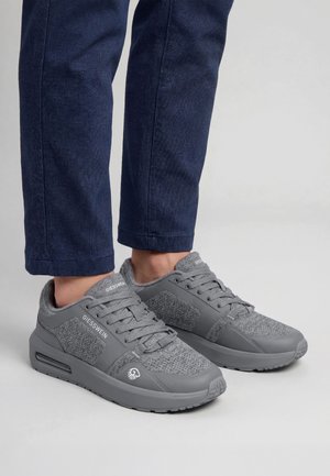 Osoba nosząca szare dziergane sneakersy Giesswein z sznurowadłami i wyściełanymi podeszwami, zestawione z ciemnoniebieskimi spodniami na jednolitym tle.