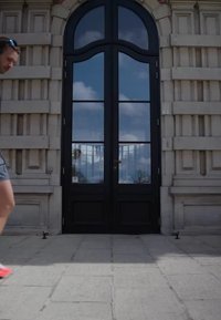 Uomo con occhiali da sole e pantaloncini che cammina davanti a una porta di vetro alta con cornice nera inserita in una facciata di pietra grigia.