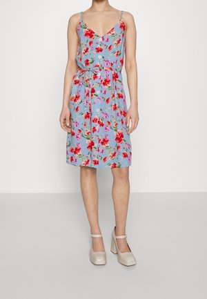 Kobieta ubrana w niebieską sukienkę na guziki bez rękawów z czerwonym i różowym kwiatowym wzorem, z talią podkreśloną paskiem, oraz beżowe buty na wysokim obcasie.