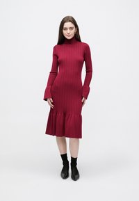 Rotes, geripptes Strickkleid mit hohem Ausschnitt, ausgestellten Ärmeln und einem gerüschten Saum. Glatte Textur mit figurbetonter Silhouette.