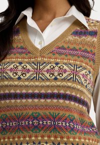 Gros plan sur un gilet en tricot multicolore avec des motifs géométriques et floraux porté sur une chemise blanche à col.