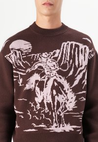 Suéter de punto marrón con un diseño de vaquero rosa y caballo contra un fondo desértico, con detalles de cactus y montañas.