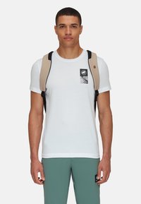Maglietta bianca in cotone con un design della tasca grafica nera. Il modello indossa bretelle beige sulle spalle, pantaloni turchesi e un'etichetta con logo.