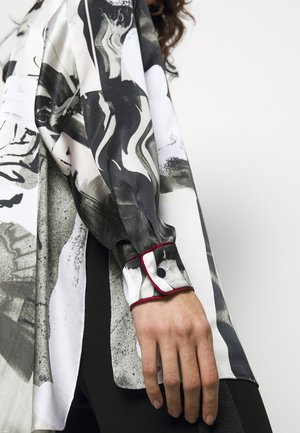 Person iført en langærmet bluse med sort, hvidt og gråt abstrakt mønster, med knaplukket manchet og røde detaljer.