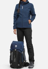 Veste softshell marine avec capuche, pantalon noir et un sac à dos bleu-noir avec des fermetures éclair et un logo, accompagné de chaussures de randonnée grises.