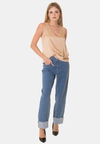 Top sans maniche in satin beige, design a scollo a V. Jeans in denim azzurro a vita media con orli arrotolati. Tacchi a punta neri. Sfondo bianco semplice.