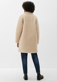 Manteau beige fluffy texturé avec col montant, manches longues et coupe droite, associé à un jean bleu foncé et des bottines noires.