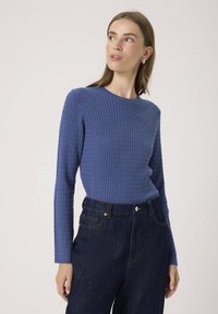 Blå ribbet langærmet top med rund hals, parret med højtafsnitne mørke denim jeans med en frontknaplukning og sidelommer.