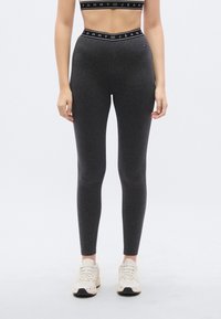 Γκρι κολάν με ψηλή μέση που διαθέτει μια αντίθετη μαύρη ταινία με λογότυπο. Το ύφασμα είναι λείο και ελαστικό, σχεδιασμένο για άνεση και εφαρμογή.