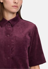 Kobieta nosząca koszulę z krótkim rękawem, zapinaną na guziki, z kieszenią na piersi, wykonaną z teksturowanej fioletowej sztruksowej tkaniny.