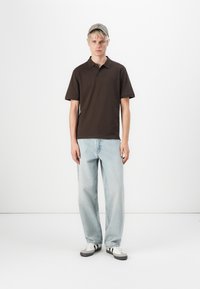 Bruine polo, lichtblauwe jeans, witte sneakers met zwarte accenten en een lichte pet. Model staat rechtop tegen een effen achtergrond.