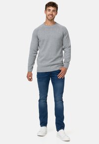 Maglione grigio lavorato a maglia con polsini e orlo a costine, abbinato a jeans blu e sneakers bianche. Il modello posa in piedi con le mani rilassate.
