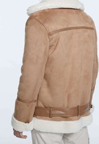 Achteraanzicht van een persoon die een lichtbruine suède jas met witte schapenvachtvoering bij de kraag, manchetten en onderrand draagt, in combinatie met lichtgekleurde broek.